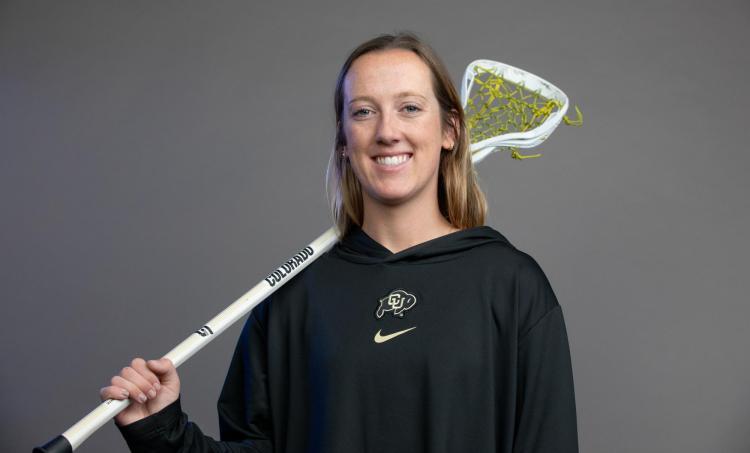 Ashley Stokes poses in a CU hoodie against a gray background while holding a lacrosse stick.
