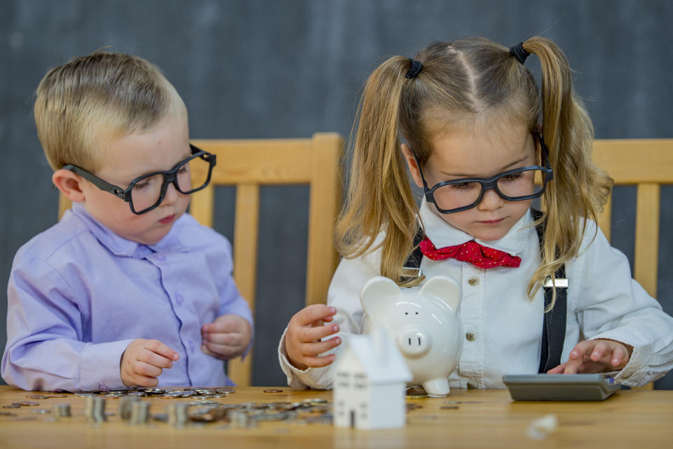 Two toddlers are wearing glasses and professional attire. They are counting money using a calculator. There is a piggy bank, piles of change, and a toy house.