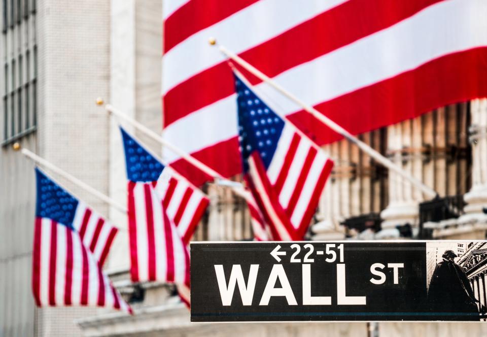 A large American flag draped over the New York Stock Exchange, with the Wall St street sign in the foreground.