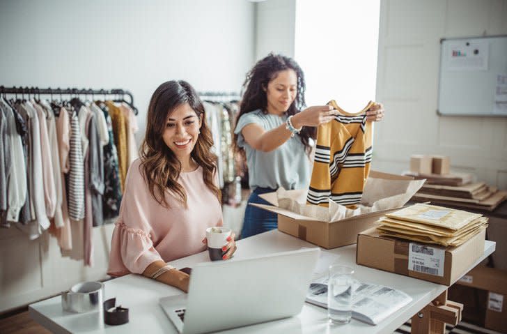 One woman packing clothes into shipping boxes while the other checks her laptop at a table in a clothing store storeroom.