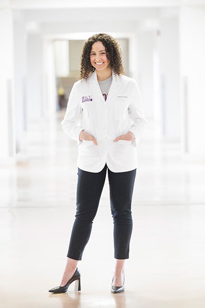 A woman medical student stands in her medical white coat and smiles against a brick background.