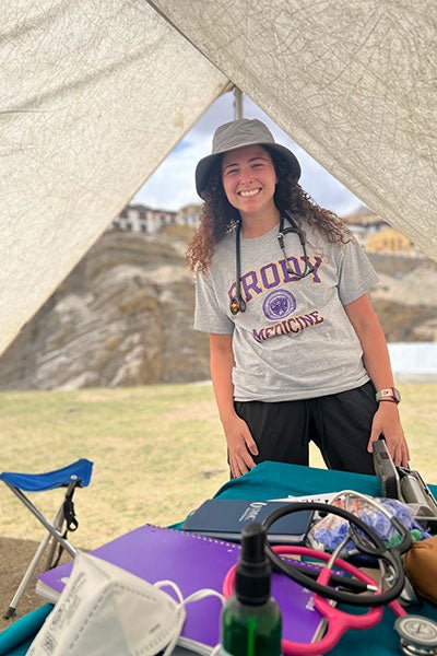 A medical student wearing a Brody School of Medicine stands at the opening of a field tent on a cliff overlooking the Himalayas in India.